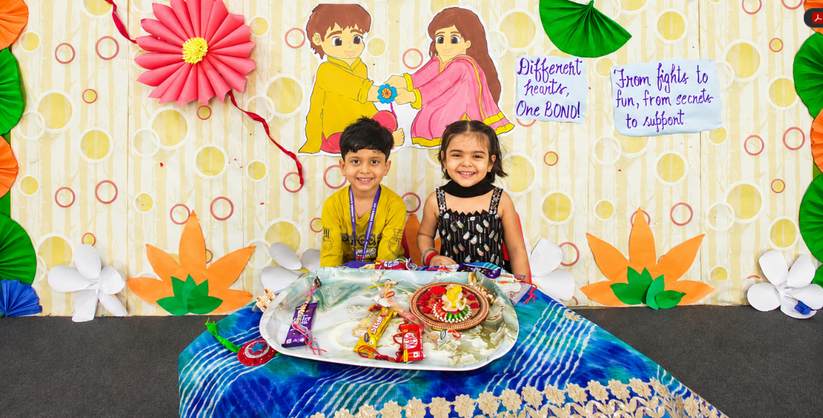 Two young students in traditional attire seated behind a decorated rakhi thali during the Raksha Bandhan celebration